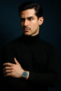 A highly stylized portrait of me with sharp features, flawless fair skin, and sleek black hair, wearing a black turtleneck sweater. natic, I’m standing against a bold navy gradient background, cofidenly crossing his arm and re showing a stylish wristwatch with blue dial and brown strap. The lighting is dramatic and cinematic, emphasizing his facial structure and giving luxury fashion magazine vibe.